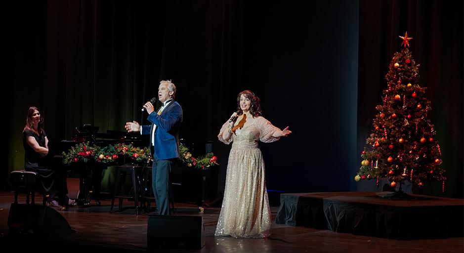 A man in a suit and a woman in a gold dress sing in front of a christmas tree accompanied by a woman playing piano. 