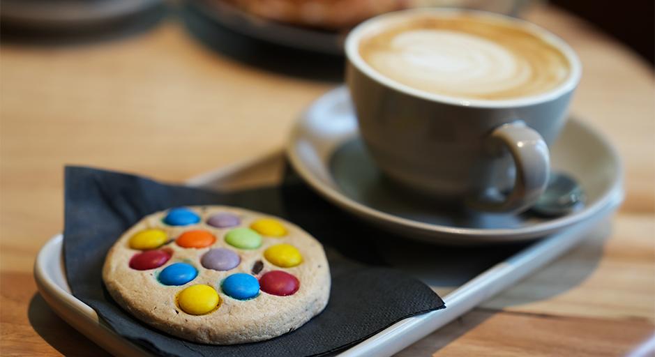 A cookie with smarties sits on a tray with a coffee beside it. 
